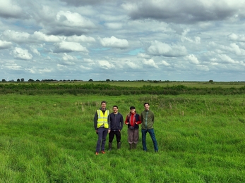 Drone image of four people standing in a field with trees on horizon and blue but very cloudy sky above