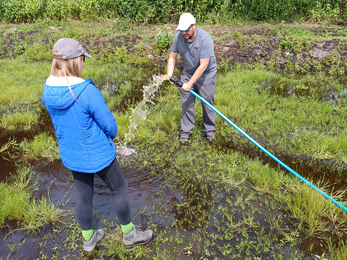 Man stands in field holding hose with running water watched by made in jacket and walking boots