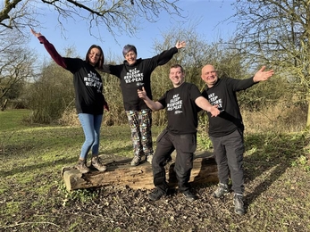 Two women stand on a log bench, two men stand in front, all arms outstretched smiling and wearing t-shirts that say Eat, Sleep, Restore, Re-Peat