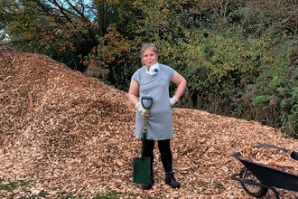 Tansey stands in front of a large pile of woodchip, holding a spade