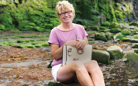 Carol sits on the beach with her sketchbook