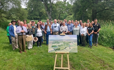 Group of 40 adults standing in a group smiling to camera, in front of them is a landscape watercolour on a wooden easel