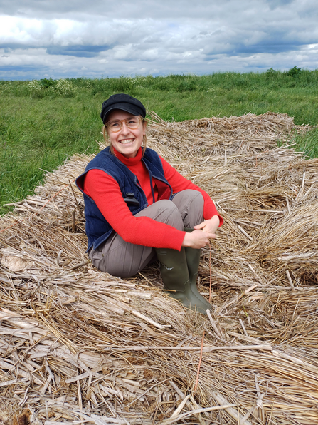 Georgemma sits on a pile of bulrush stems in a field. She smiles off camera and is wearing glasses, a red polo-neck jumper, blue waistcoat and hat