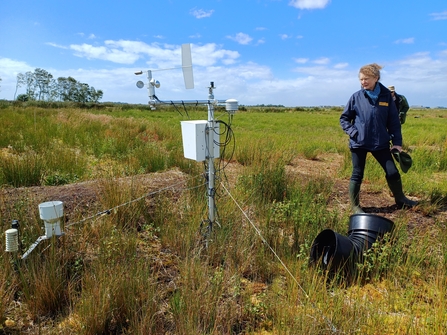 Kate stands in blue jacket and wellies looking at monitoring equipment places in the ground in a field, blue sky overhead