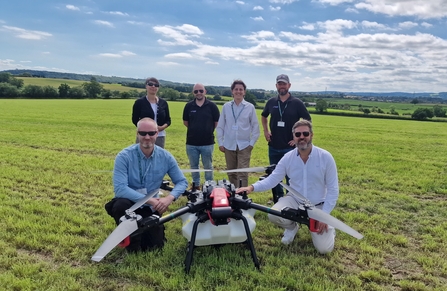 Four people stand and one kneels either side of a large drone in a field on a sunny day