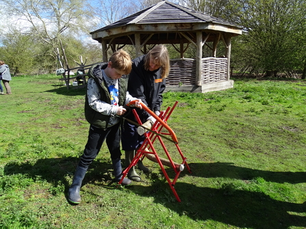 A boy uses a bow saw to cut a log on a saw horse, supported by an adult volunteer