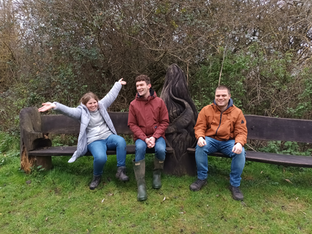 Tansy, Lewis and their support worker sit on a wooden bench featuring a carved great crested newt. 